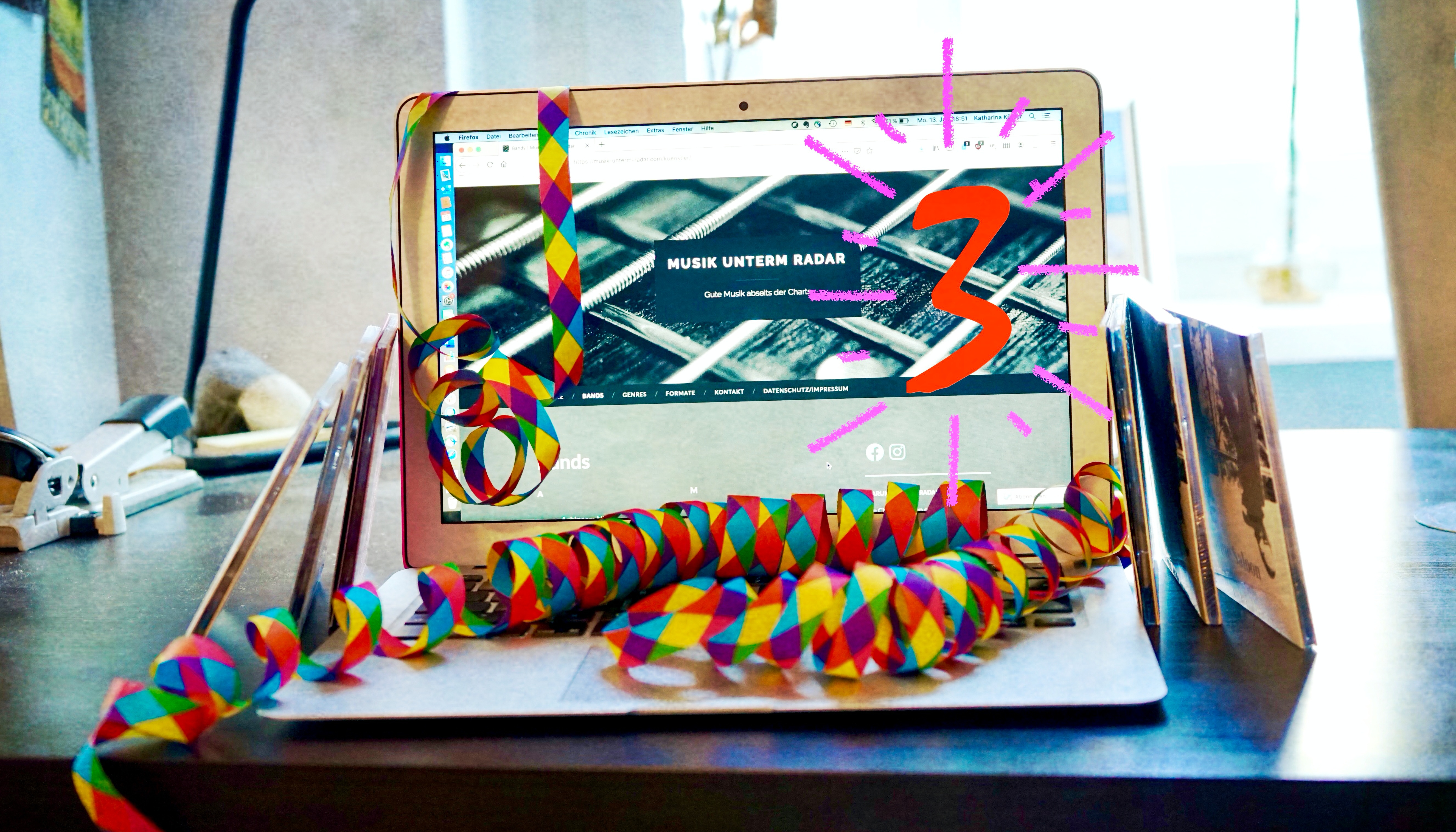 Laptop mit Luftschlangen und sechs CDs. Der Bildschirm zeigt die Website Musik unterm Radar und die Zahl drei zum dreijährigen Jubiläum.