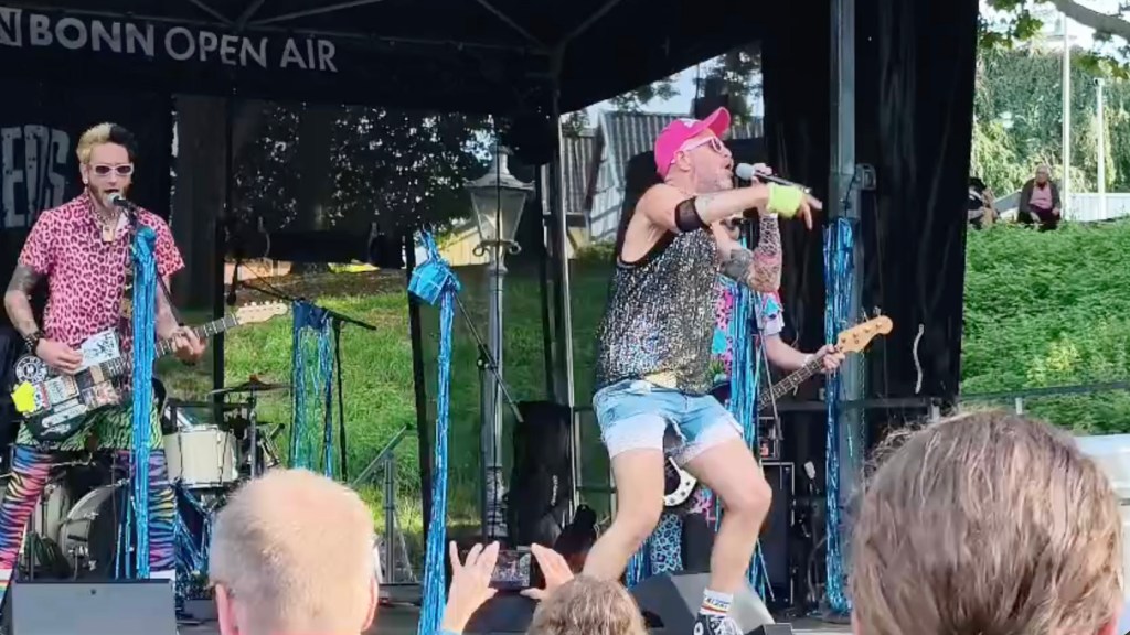 Ein energiegeladener Auftritt einer Band auf der Bühne beim Bonn Open Air, mit einem Sänger in Glitzeroberteil und kurzen Hosen, umgeben von weiteren Bandmitgliedern in bunten Outfits.